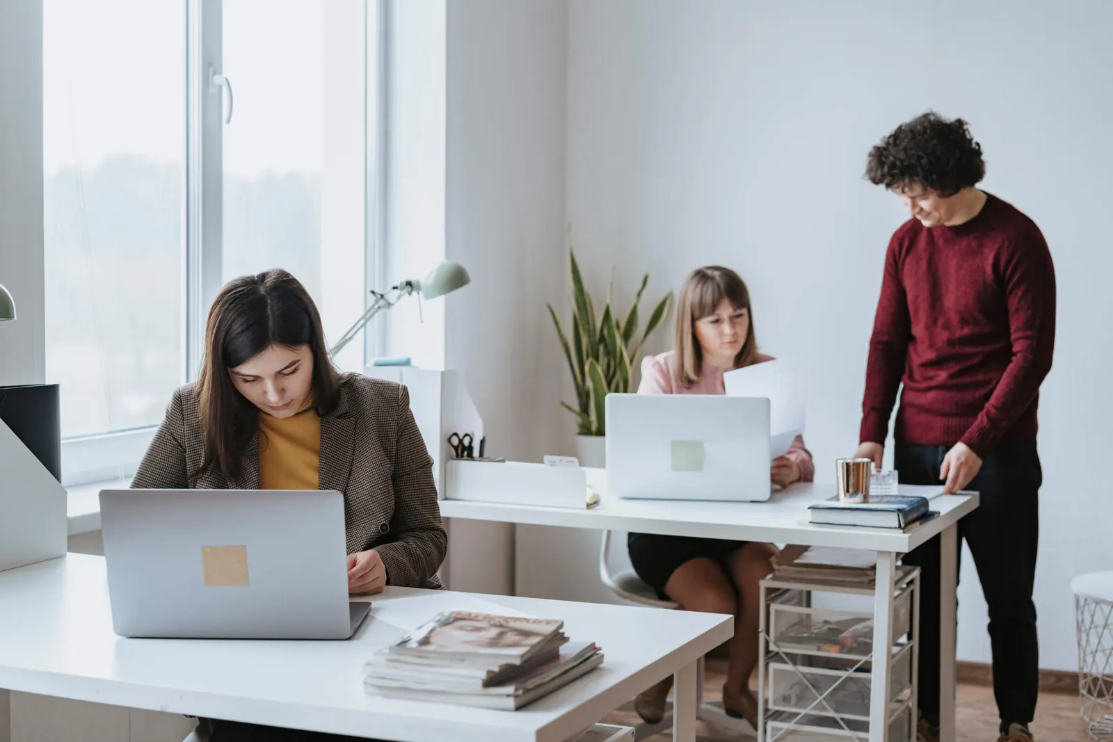 Tiga rekan kerja di kantor yang terang: dua bekerja di laptop pada meja terpisah dan satu berdiri membaca dokumen.