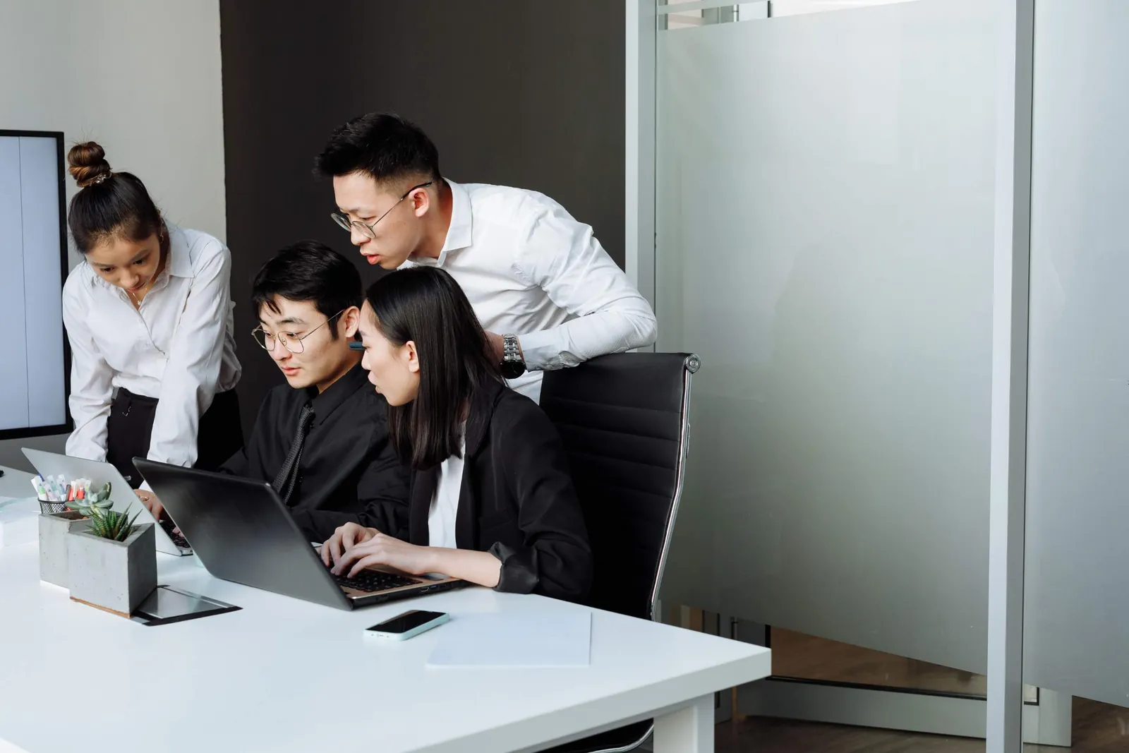 Four colleagues gathered around a laptop in a modern office meeting room, two seated and two leaning in to look at the screen.