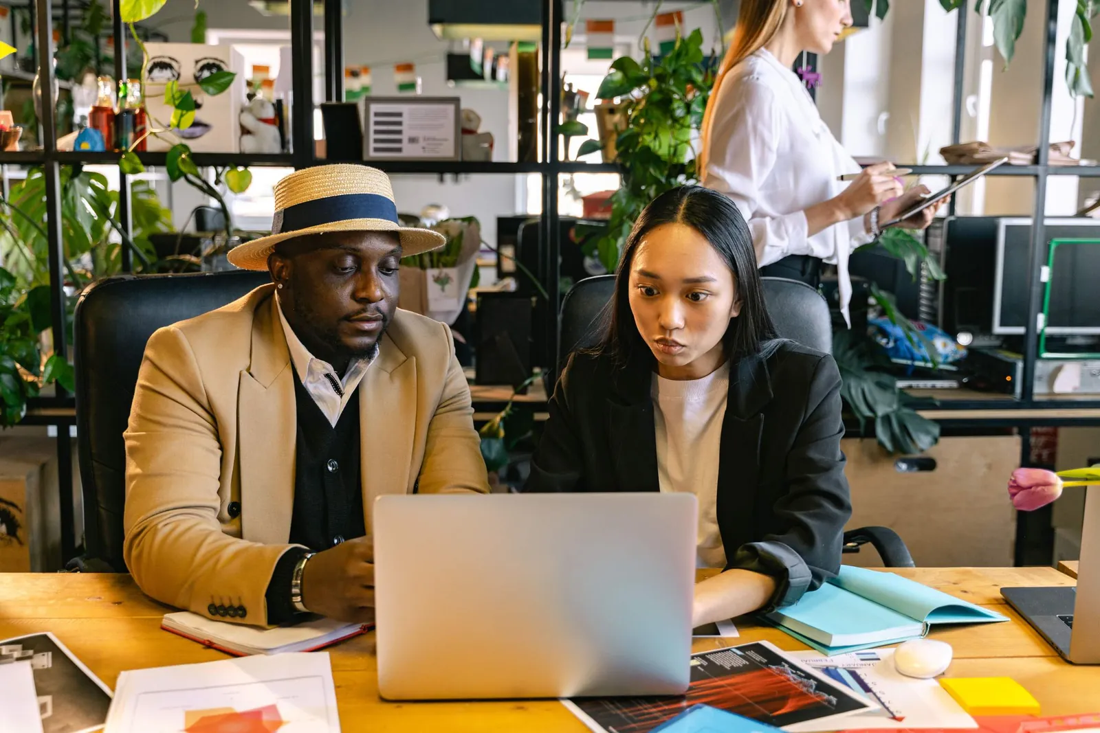Dua rekan kerja duduk berdampingan di meja bersama, menatap laptop dengan saksama, sementara rekan ketiga bekerja dengan tablet di belakang mereka di kantor penuh tanaman.
