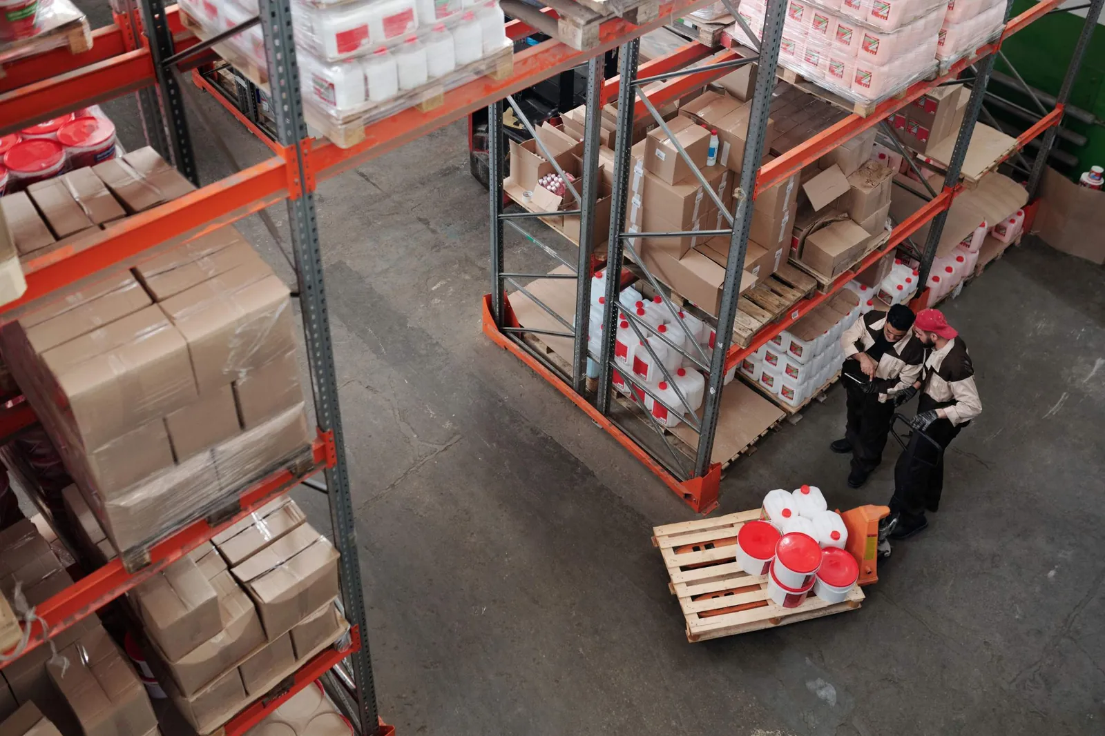 Dua pekerja gudang berseragam memeriksa tablet di samping pallet jack berisi wadah, dikelilingi rak tinggi berisi stok dalam kardus.