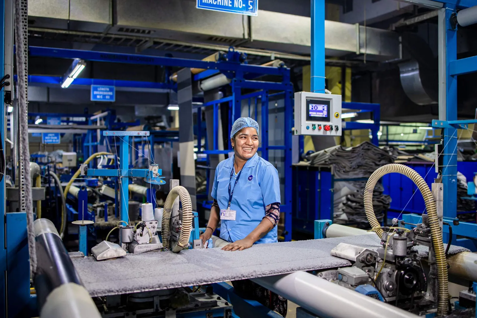 A smiling production worker in a blue uniform and hairnet stands at a machine in a factory hall.